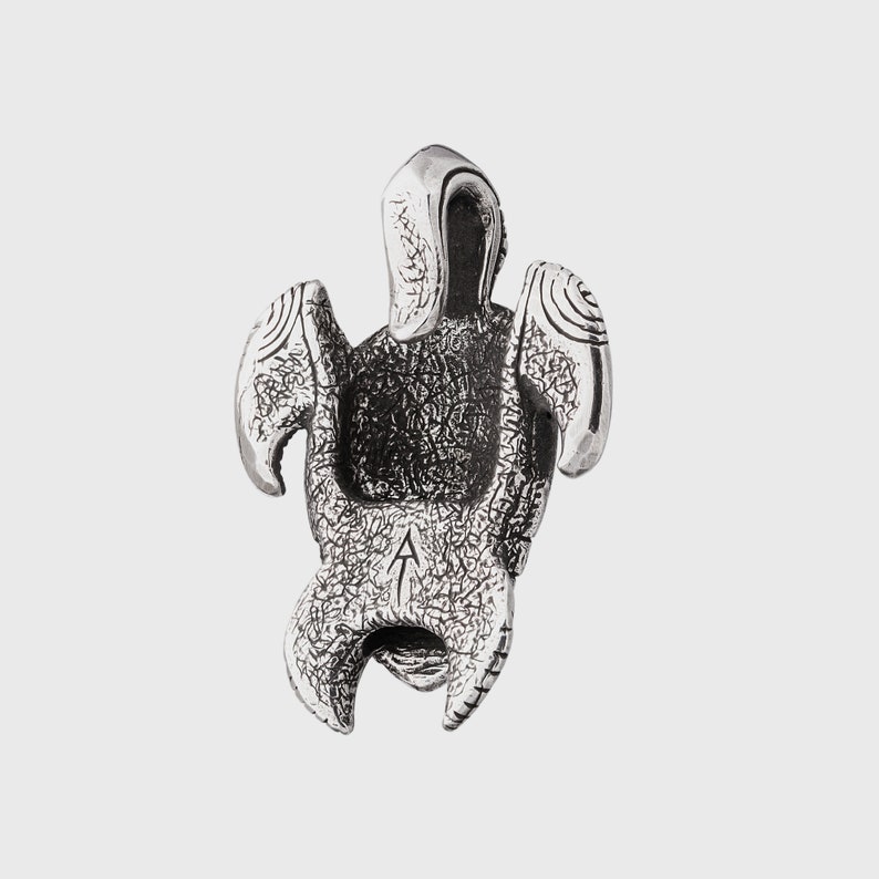 May include: A silver pendant shaped like a turtle with textured detailing. The pendant features wing-like elements and a curved head. A small letter "A" is engraved on the bottom. The pendant is likely made of a metal alloy.