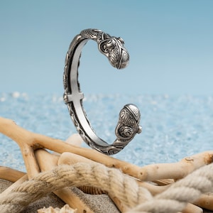 Peut inclure: Bracelet manchette argenté avec des gravures ornées et détaillées. Le bracelet présente des extrémités décoratives et une fermeture à charnière. Le fond comprend du bois flotté et un fond bleu semblable à l'océan.