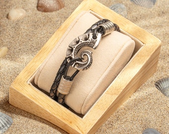 May include: A black leather bracelet with a silver hook clasp. The hook is decorated with a detailed design. The bracelet is displayed in a wooden box on a sandy surface.