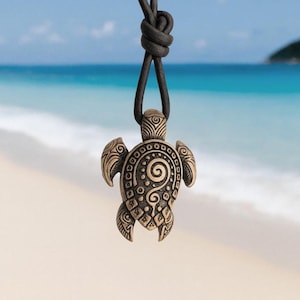May include: A bronze turtle pendant necklace with a black cord. The turtle pendant has detailed engravings, including a spiral design on its shell. The necklace is shown against a blurred beach background with blue sea and sky.