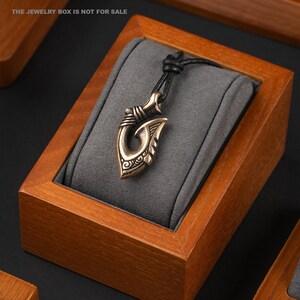 May include: A bronze pendant in the shape of a fish hook with intricate details hangs from a black leather cord. The pendant is displayed in a wooden box with a gray velvet lining. "The Jewelry Box is not for sale" is printed on the box.