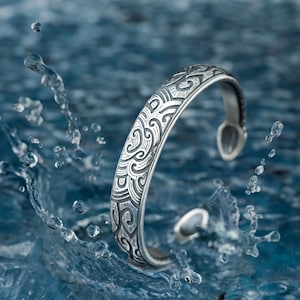 May include: A silver bracelet with an intricate wave pattern design. The bracelet is set against a blue water background.