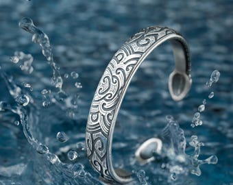 May include: A silver bracelet with an intricate wave pattern design. The bracelet is set against a blue water background.