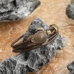 Könnte beinhalten: Ein braunes Lederarmband mit einem bronzenen Schildkrötenanhänger. Der Schildkrötenanhänger hat aufwendige Details und ein Spiralmuster auf seinem Panzer. Das Armband wird auf einem grauen Felsen präsentiert und unterstreicht seine erdigen Töne und sein nautisches Thema.