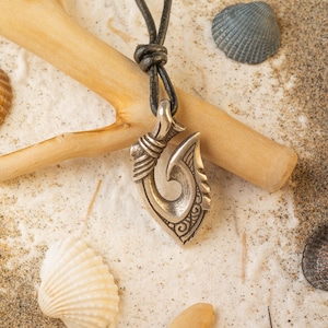 Peut inclure: Un collier avec un pendentif en forme d'hameçon de pêche en argent sur un cordon en cuir noir. Le pendentif est orné de sculptures complexes et est présenté sur une surface de sable blanc avec des coquillages.
