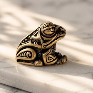 Könnte beinhalten: Eine kleine Froschfigur aus goldfarbenem Metall mit komplizierten, geprägten Mustern auf dem Körper. Der Frosch ist in sitzender Position dargestellt und weist eine strukturierte Oberfläche, markante Augen und stilisierte Zeichnungen auf.