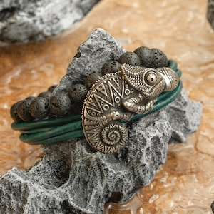 May include: A green leather bracelet with black lava rock beads and a silver chameleon charm. The charm is detailed with intricate patterns and has a raised eye.