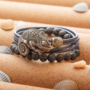 Op de afbeelding: Een blauwe leren armband met zwarte lavastenen kralen en een zilveren kameleon bedel. De armband is gelaagd met meerdere rijen leer.