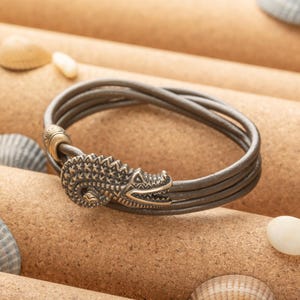 Peut inclure: Un bracelet en cuir marron avec un fermoir en forme de crocodile. Le bracelet est composé de trois brins de cuir et possède une perle décorative près du fermoir.