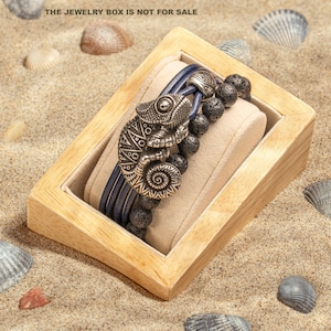 May include: A blue and silver bracelet with a chameleon charm, displayed in a wooden box on a sandy surface. The jewelry box is not for sale.