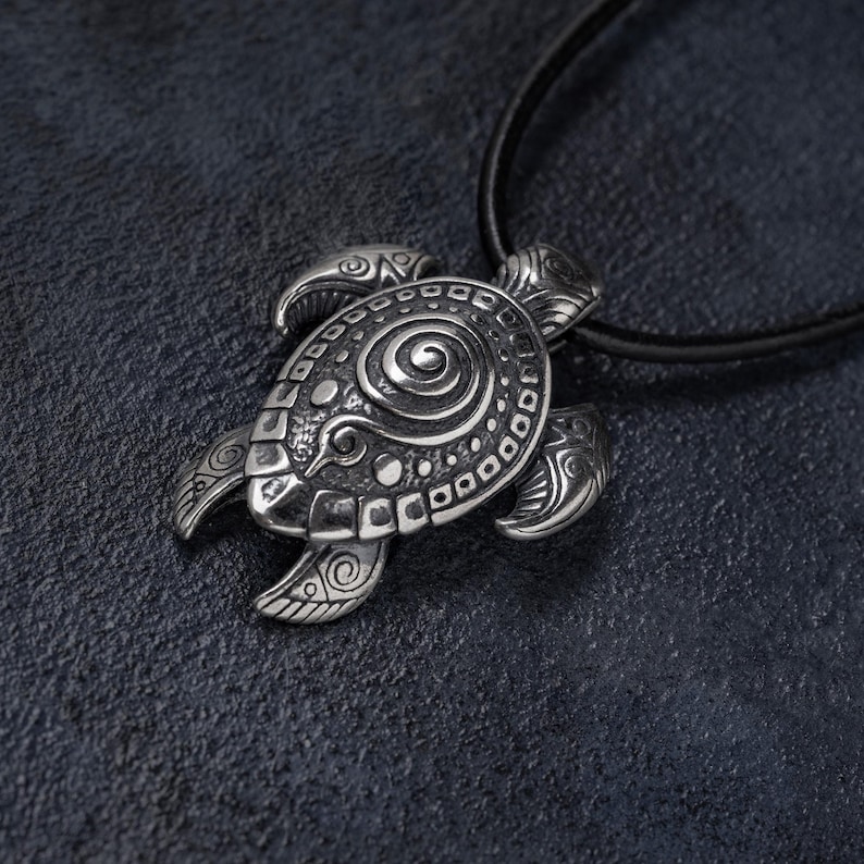 May include: Silver turtle pendant necklace. The pendant has detailed patterns and a spiral design on the shell. The necklace has a black cord. The turtle pendant is a symbol of longevity and wisdom.