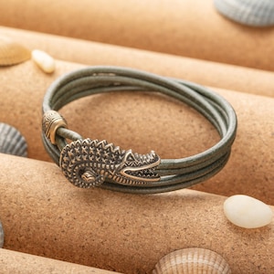 May include: A green leather bracelet with a decorative alligator clasp. The bracelet is made of three strands of leather and has a textured finish.