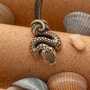 May include: A bronze snake pendant with intricate details, hanging on a black leather cord. The snake is coiled and has a detailed head with a textured surface.