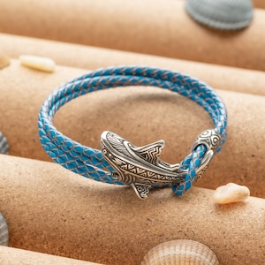 Może przedstawiać: Bransoletka ze skóry w kolorze niebieskim z zawieszką w kształcie rekina ze srebra. Bransoletka jest pleciona i ma srebrną klamrę.