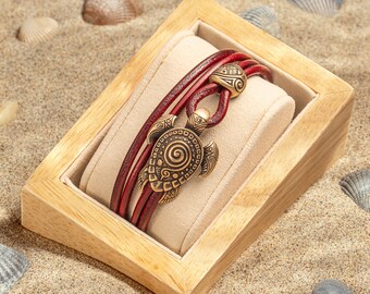 May include: A red leather bracelet with a gold turtle charm. The bracelet is displayed in a wooden box on a sandy surface.