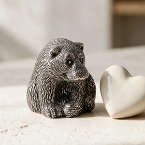 Pode incluir: Uma pequena estatueta de urso cinza-escuro texturizada com marcas de pelo detalhadas, posicionada ao lado de um objeto liso em forma de coração na cor creme. O urso é retratado em uma postura sentada com a cabeça levemente virada.