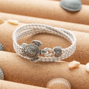 Könnte beinhalten: Weißes Lederarmband mit einem silbernen Schildkrötenanhänger. Das Armband ist geflochten und hat eine dekorative Perle.