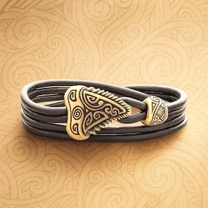 May include: A black leather bracelet with a gold-colored clasp and bead. The clasp features an intricate design, and the bracelet has multiple strands. The bead also has a detailed pattern.