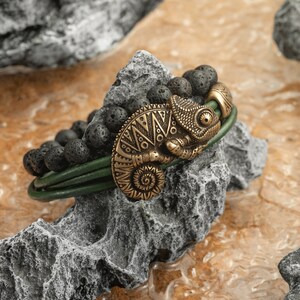 Op de afbeelding: Een groene leren armband met zwarte lava kralen en een bronzen kameleon bedel. De armband ligt op een grijze rots achtergrond.