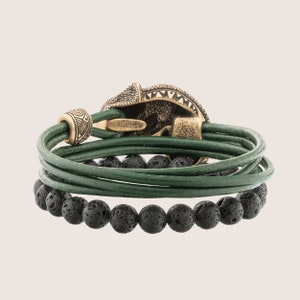 Op de afbeelding: Een groene leren armband met een zwarte lavastenen kralenaccent en een gouden metalen sluiting. De sluiting heeft een gedetailleerd ontwerp.