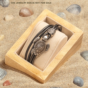 Pode incluir: Uma pulseira de couro preto de várias camadas com um pingente de metal prateado em forma de tartaruga marinha com um padrão de espiral na carapaça e uma conta cilíndrica a condizer, sobre uma almofada de veludo bege numa caixa de madeira clara.