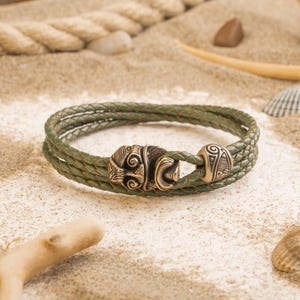 Könnte beinhalten: Geflochtenes Armband aus olivgrünem Leder mit dekorativem antikgoldfarbenem Verschluss. Das Armband besteht aus mehreren Ledersträngen und weist auf dem Verschluss aufwendige Details auf. Ein stilvolles Accessoire.