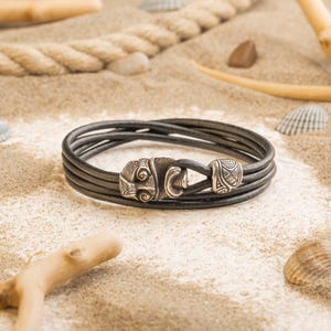 Könnte beinhalten: Ein schwarzes Lederarmband mit einem silbernen Verschluss. Das Armband hat mehrere Stränge und liegt auf einer sandigen Oberfläche mit Muscheln und Treibholz.