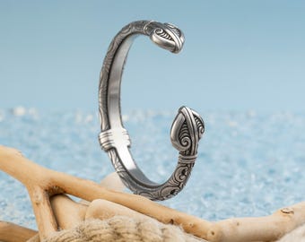 May include: A silver-toned cuff bracelet with ornate, swirling designs. The open-ended bracelet features decorative ends and detailed patterns along its curved surface. The bracelet is displayed on a beach-themed background with driftwood and rope.