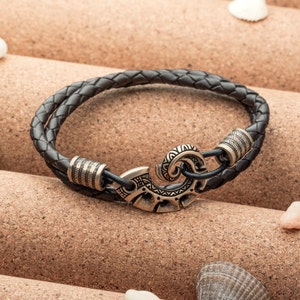 May include: Braided black leather bracelet with a decorative clasp. The bracelet features a double-strand design and antique gold-colored metal accents. The clasp has an intricate, stylized design. A stylish accessory.
