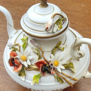 Op de afbeelding: Witte keramische theepot met gouden details, waaronder een handvat, tuit en accenten. Versierd met kleurrijke, driedimensionale bloemen en bladeren. Het deksel heeft een gouden knop.