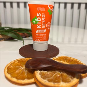 May include: Orange tube of Wooden Spoon Kids Toothpaste with the text "KIDS TOOTHPASTE" and a green cartoon character. The tube is standing on a brown coaster, with dried orange slices and a wooden spoon in the foreground.
