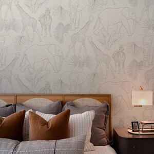 May include: A bedroom with a bed featuring a headboard with a woven design and a variety of pillows in shades of brown and gray. The wall behind the bed has a white wallpaper with a black line drawing pattern of animals, including giraffes, elephants, and zebras. A nightstand with a lamp and a tray of framed photos is next to the bed.