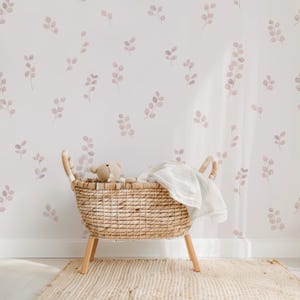 May include: A woven wicker basket with wooden legs, holding a teddy bear and a white blanket, sits on a beige rug. The background is a white wall decorated with a pattern of pink leaf sprigs.