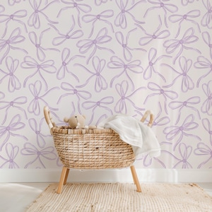 May include: A white wall with a repeating pattern of light purple watercolor bows. A wicker basket with a white blanket and a stuffed animal sits in front of the wall.