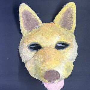 May include: A handmade dog mask with yellow and brown coloring. The mask features large, upright ears, a brown nose, and a pink tongue. The eye holes are cut out, and the mask appears to be made of a textured material.