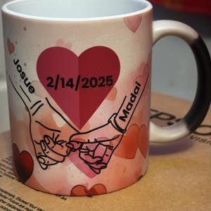 May include: A white ceramic mug with a black handle. The mug has a pink and red watercolor design with two hands holding pinkies, a large red heart with the text "2/14/2025" and the names "Josue" and "Madai" written on either side of the heart.