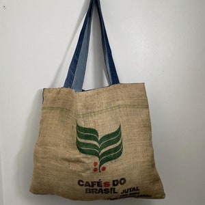 Puede incluir: Una bolsa de lona marrón con asas de mezclilla. La bolsa tiene un diseño de hoja verde y el texto "CAFÉS DO BRASIL JUTAL 100% CARBON" impreso en la parte delantera.