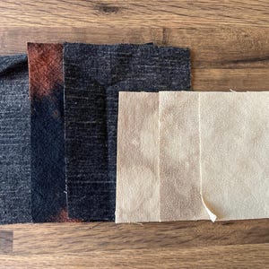 Puede incluir: Una colección de muestras de tela en varios colores y texturas. Las muestras incluyen gris oscuro, negro con un degradado marrón rojizo y tonos beige. Las telas están dispuestas sobre una superficie de madera, mostrando sus diferentes tejidos y tonos.