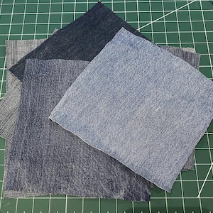 May include: Three squares of denim fabric in different shades of blue, arranged in a stack on a green cutting mat.
