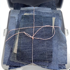 Può includere: Un mucchio di quadrati di tessuto denim legati insieme con uno spago rosa. Il tessuto è di diverse tonalità di blu.