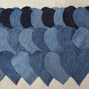 Puede incluir: Colección de corazones de mezclilla en varios tonos de azul, dispuestos juntos. Los corazones están cortados de tela de mezclilla, mostrando la textura y las variaciones de color del material. El fondo es de color beige neutro.