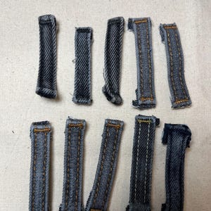May include: Ten denim belt loops in various stages of completion. The loops are a medium blue with visible stitching in a contrasting tan colour. The fabric has a textured appearance, and the loops are arranged on a light-coloured fabric surface.