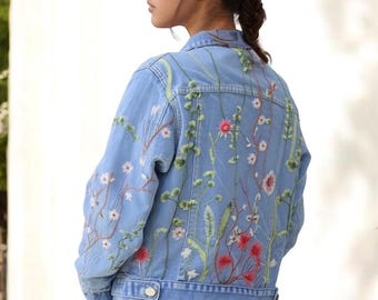 May include: Light blue denim jacket with embroidered floral design in red, white, and green. The jacket features a classic collar, button closure, and long sleeves. The back of the jacket is adorned with a detailed floral pattern. The model is wearing a matching denim skirt.