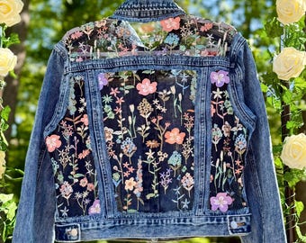 May include: A blue denim jacket with a floral embroidered sheer panel on the back. The embroidery is in shades of pink, purple, orange, and green.