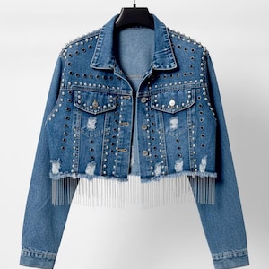 May include: A medium-wash blue denim jacket featuring a cropped silhouette, distressed frayed hem, and long sleeves. The jacket is embellished with silver-toned metal studs and black circular accents, finished with a long silver chain fringe hem.