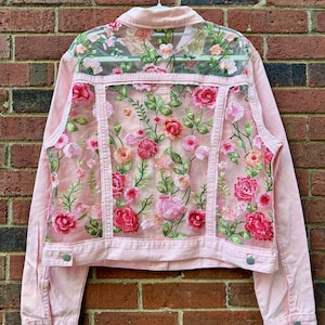 Op de afbeelding: Een lichtroze spijkerjas met een transparant mesh-achterpand, versierd met gedetailleerd bloemenborduursel in rood-, roze- en groentinten. De jas is voorzien van een klassieke kraag, lange mouwen met knoopmanchetten en zilverkleurige knopen.
