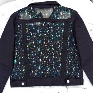 May include: Black denim jacket with a sheer panel on the upper back and a star pattern. The star pattern is a mix of iridescent colors. The jacket has two front pockets and two buttons on the front.