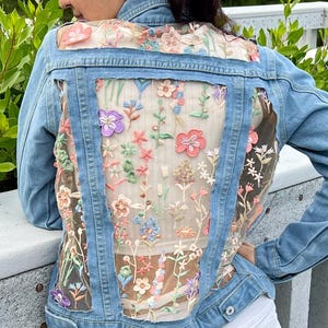 以下が含まれることがあります： 薄いブルーのデニムジャケットで、背面に透き通るような花の刺繍のパネルが付いています。刺繍はピンク、パープル、グリーン、ホワイトの色合いです。