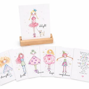 May include: A set of illustrated greeting cards featuring whimsical drawings of girls with inspirational words like "strength" and "brave." One card is displayed in a wooden holder, showcasing a girl with a dog. The cards have a watercolor style.