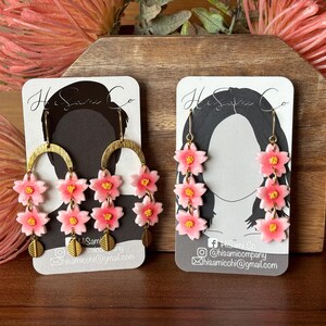 May include: A pair of pink flower dangle earrings with gold hoops and a gold chain. The earrings are displayed on a white card with the text "Hi Sam Co." and contact information.